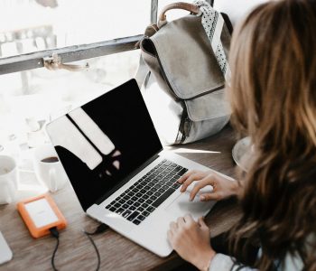 A young woman works remotely at a café, using her laptop and external hard drive.